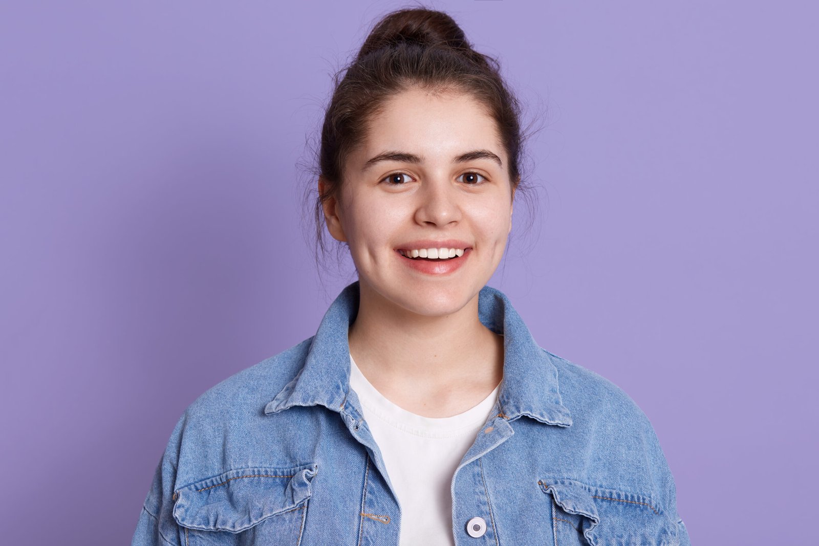 Smiling happy woman wearing denim jacket and white shirt, looking directly at camera with toothy smile, lady with hair bun, posing isolated over lilac background.