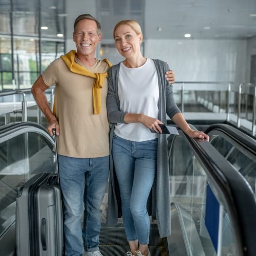 Full-length portrait of a smiling male passenger with the trolley suitcase hugging his female companion on the moving staircase