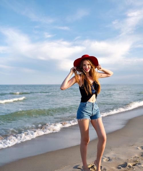 Summer fashion portrait of young woman walking alone near ocean, vacation on the beach, travel alone, wearing vintage hat sunglasses and denim shorts, slim body, sunrise, healthy lifestyle.